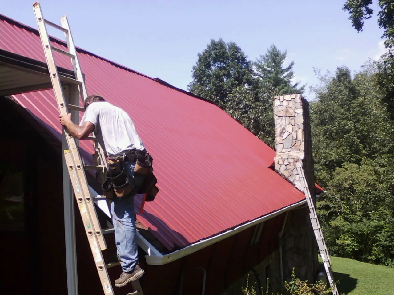 Roofer on a ladder beside a red metal roof and stone chimney — Soffit & Fascia Repair by Roofing Brothers in East Wenatchee